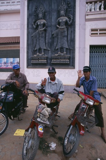 CAMBODIA, Siem Reap, Three motorbike taxi drivers on their mopeds beneath a large wall plaque depicting Asparas