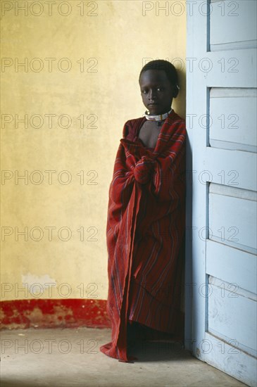 TANZANIA, Ngorongoro, Masai child standing between yellow wall and blue door.  The Masai have been expelled from the Ngorongoro Crater area to make way for tourists and wildlife.