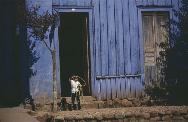 CHILE, Urban Scene, Young child standing in doorway of blue painted building swinging lasso.