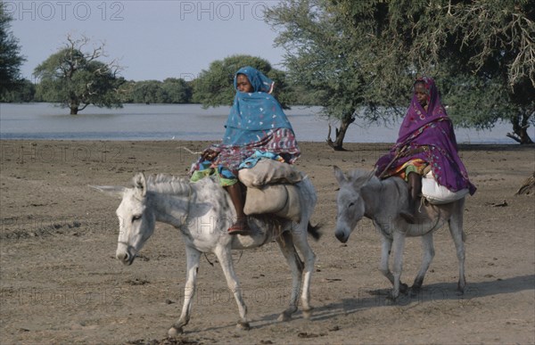 SUDAN, South Darfur, Transport, Baggara Arabs.  Two women from the Beni Halba tribe riding donkeys past lake.