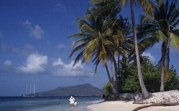 WEST INDIES, Grenadines, Carriacou Island, Sandy Island.  Tourists on sandy beach beneath palm trees with sailing ship moored in water beyond.