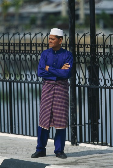 BRUNEI, People, Men, Man standing on pavement with arms crossed next to railings dressed up to visit the Omar Ali Saifuddin Mosque in Bandar Seri Begawan.