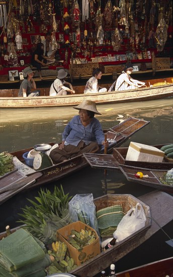 THAILAND, South, Bangkok, Damnoen Saduak Floating Market with an old female fruit vendor in her canoe and tourists in a canoe behind in front of a tourist goods stall