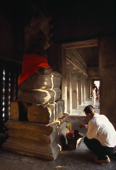 CAMBODIA, Siem Reap Province, Angkor Wat, Interior of shrine on the upper level with visitor lighting incense at base of Buddha figure seated below naga with multi headed hood.