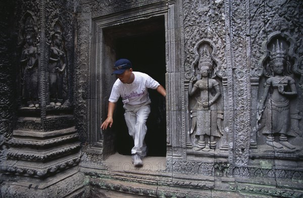 CAMBODIA, Siem Reap Province, Angkor, Ta Prohm.  Visitor climbing through doorway of twelth century Buddhist temple with exterior wall decorated with bas relief carvings.
