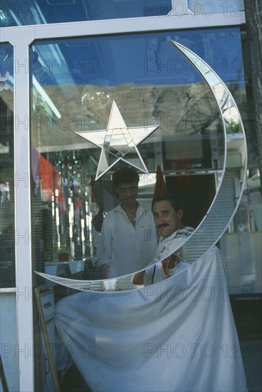 PAKISTAN, Northern Areas, Gilgit, Barbers shop with star and moon of the Pakistan flag inlaid on the window in mirror glass reflecting the photographer.  Customer and barber seen inside