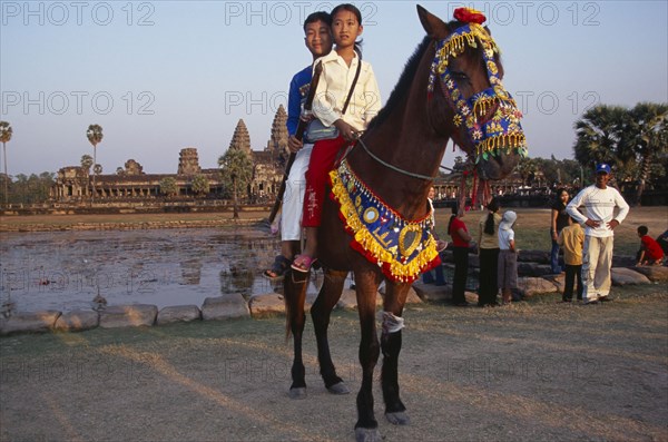 CAMBODIA, Siem Reap Province, Angkor Wat, Cambodian children visiting Angkor during the Chinese New Year having photographs taken on pony wearing decorative harness in front of lake and temple complex.