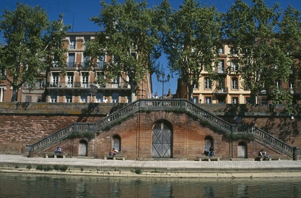 FRANCE, Midi Pyrenees, Haute Garonne, Toulouse.  Red brick steps leading down to the River Garonne from street level with facades of buildings partly seen through trees behind.  People sitting in the sun.