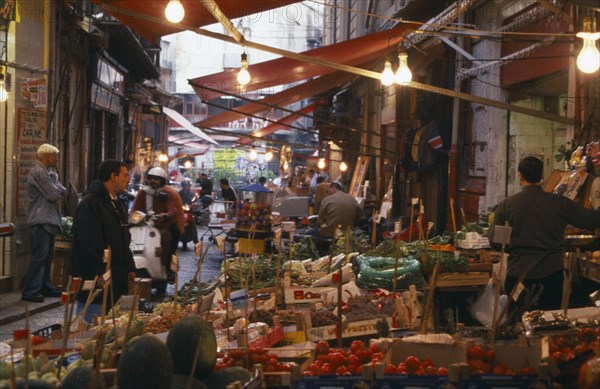 ITALY, Sicily, Palermo, Vucciria Market.  Narrow street lined with fruit and vegetable stalls.  Passing pedestrians and moped riders.