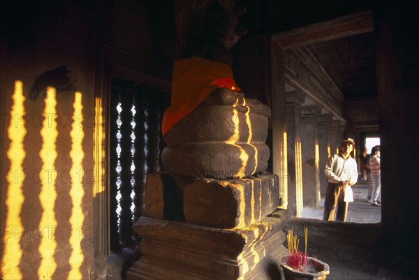 CAMBODIA, Siem Reap Province, Angkor Wat, Tourist looking into interior of shrine in upper or third level with Buddha figure seated below a naga and incense offerings buring at base.