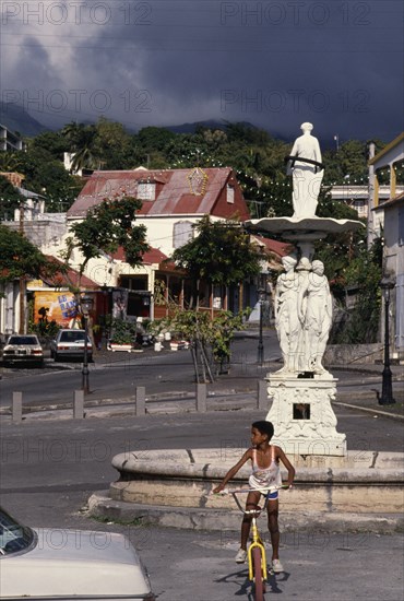 WEST INDIES, Guadeloupe, Basseterre, Large white statue in middle of street surrounded by houses and a boy on a bike in the foreground.
