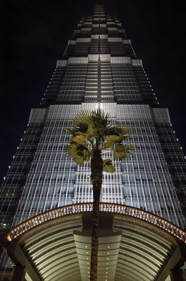 CHINA, Shanghai, Angled view looking up at the Hyatt Hotel skyscraper illuminated at dusk with entrance arch and palm in the foreground