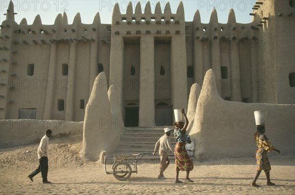 MALI, Djenne, Exterior of mud brick mosque built in traditional style with woman walking past carrying buckets on their heads.