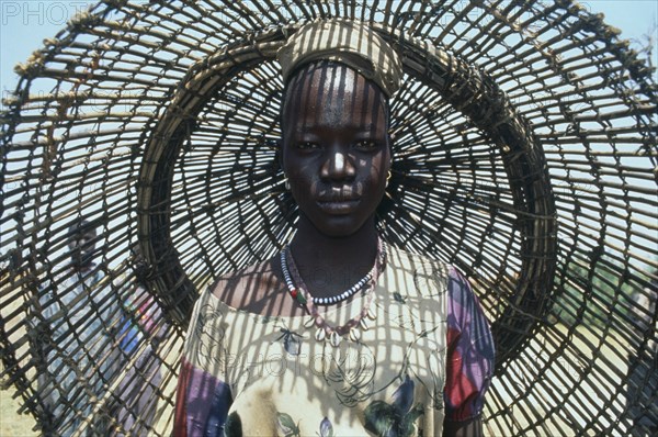 SUDAN, Bahr el Ghazal, Portrait of young Dinka woman with fishing basket behind her casting shadow pattern over her face and upper body.