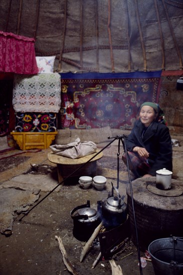 CHINA, Xinjiang Province, Altai Region, Kazakh woman inside her Kigizuy or yurt with covered stove and kettle suspended over small open wood fire.