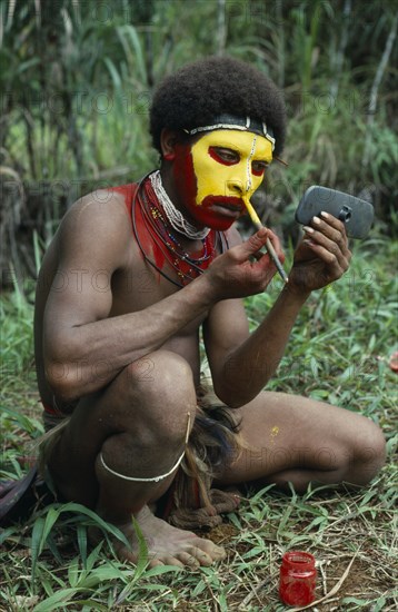 PAPUA NEW GUINEA, Southern Highlands, Tari Valley, Huli tribesman painting his face using old car wing mirror to see his reflection.
