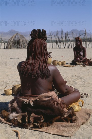 NAMIBIA, Skeleton Coast, Hoarusib Valley, Kaokoland.  Back of seated Himba woman wearing calfskins and with her body creamed with mixture of scented butterfat and ochre to give red glow.