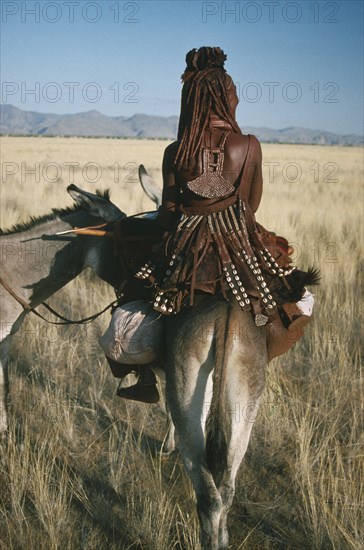 NAMIBIA, Marienfluss, Himba tribeswoman on donkey seen from behind wearing typical leatherwork decorated with cowrie shells and copper.