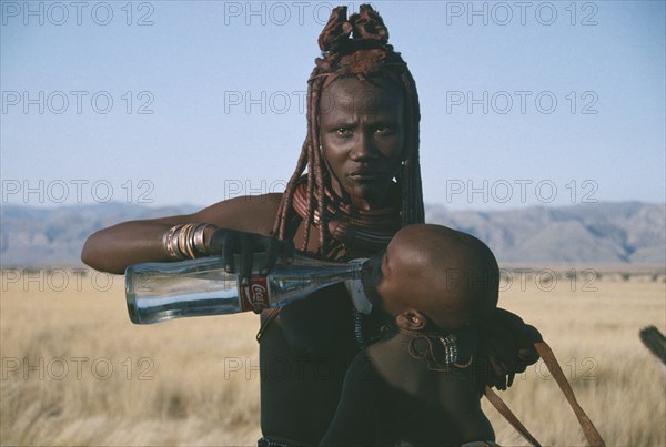 NAMIBIA, Marienfluss, Himba woman giving child a drink of water from Coca Cola bottle.  The Himba traditional way of life is being threatened by uncontrolled tourism.