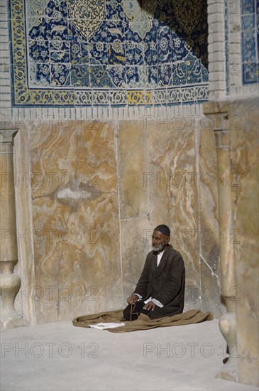 IRAN, Esfahan, Man with prayer beads praying beside wall of mosque.