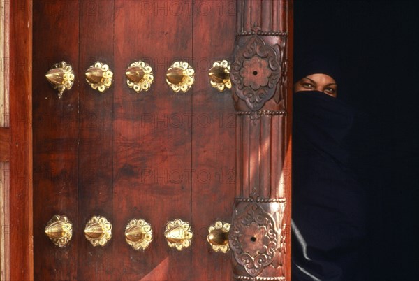 TANZANIA, Zanzibar Island, Zanzibar, Woman in full chandor looking out from behind ornate carved door.