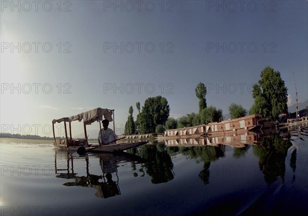 INDIA, Kashmir, Srinagar, Nagin Lake. Man in Shikara or Water Taxi with two modern houseboats moored at the edge of the lake