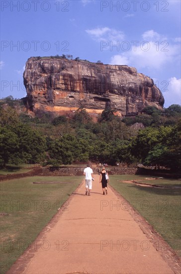 SRI LANKA, Sigiriya, View towards huge monolithic rock site of fith century citadel.  Also called Lion Rock.  Tourist couple on path through gardens in the foreground.