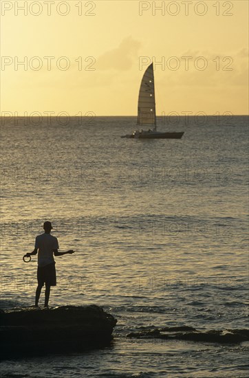 WEST INDIES, Barbados, Bridgetown, Fisherman silhouetted in the evening light with yacht on the horizon