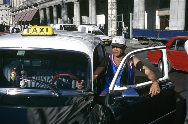 CUBA, Havana, Driver standing by the open door of his classic American style black cab parked opposite the Capitolio Building