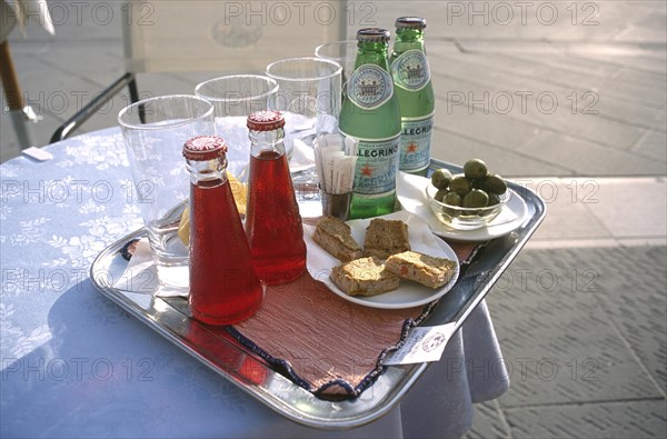 ITALY, Trieste, Piazza Unita, Tray on a small round lace clothed table with glasses and two bottles of Mineral water standing with two bottles of Campari and a dish of Olives