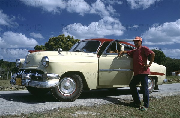 CUBA, Cienfuegos, Man standing by the open door of a classic style American car which has been fitted with tractor tyres due to a tyre shortage