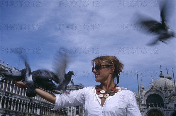 ITALY, Veneto, Venice, St Marks Square. Woman wearing a white shirt standing with pigeons fluttering in motion blur