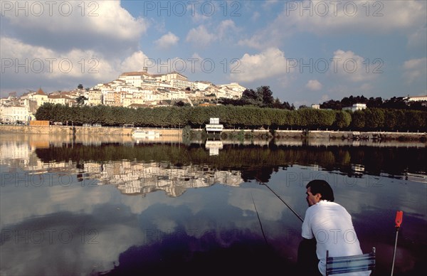 PORTUGAL, Coimbra, Man fishing on the bank of the River Mondego with the University crowning Alcacova Hill opposite