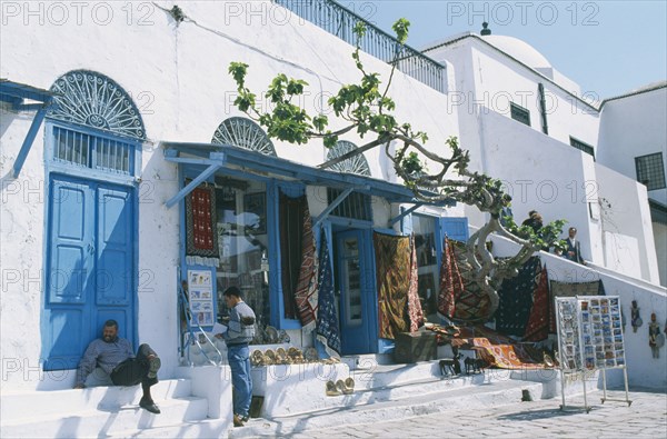 TUNISIA, Sidi Bou Said, Rugs and postcards for sale.  White painted shopfront with blue doors.  Two local men in shade of awning.
