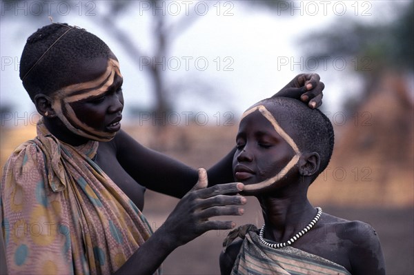 SUDAN, Tribal People, Dinka girl decorating the face of a friend using dung ash mixed with water or cattle urine.
