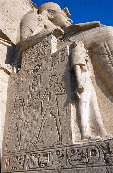 EGYPT, Nile Valley, Abu Simbel, Sun Temple of Ramses II.  Part view of one of the colossi of Ramses II flanking entrance to temple showing wall with relief carving and hieroglyphics.