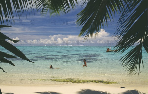 PACIFIC ISLANDS, Polynesia, Society Islands, Moorea.  Beach and swimmers partly framed by palm tree fronds.