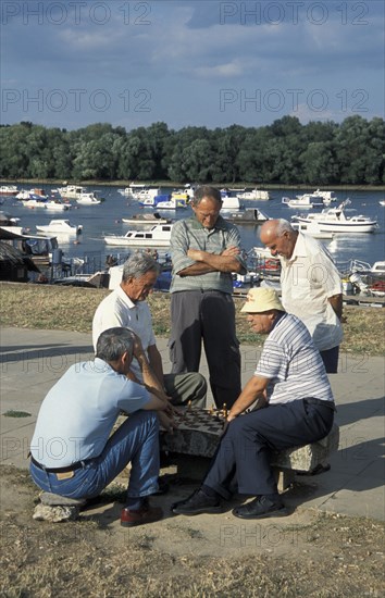 SERBIA, Belgrade, Group of men gathered around playing Chess with boats on The Danube river behind.
