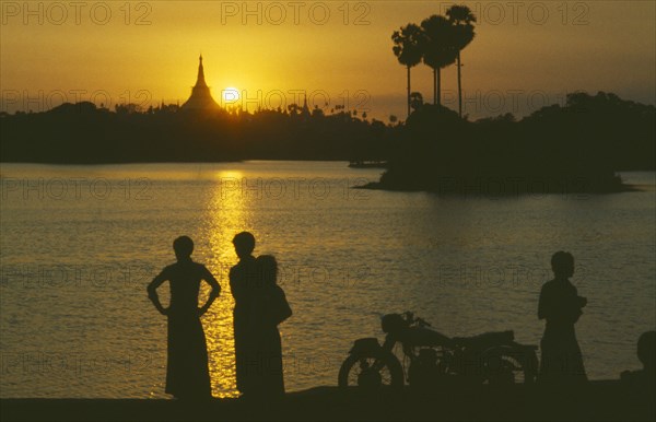 MYANMAR, Yangon, People silhouetted on shore of lake looking across towards the Shwedagon Pagoda at sunset.