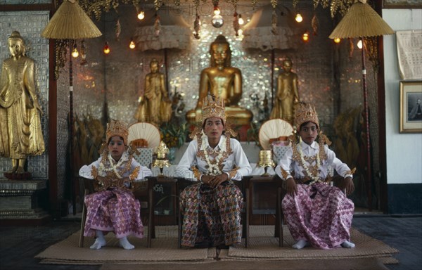 MYANMAR, Religion, Buddhism, Shwedagon Pagoda.  Novice monks at Shinpyu initiation ceremony seated in front of seated and standing golden Buddha figures.