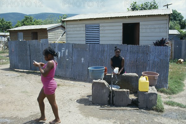 WEST INDIES, Jamaica, Kingston, Bower Bank Transit Camp.  Girls beside water pipe with simple housing behind.