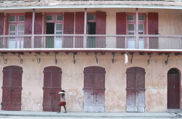 HAITI, Cap Haitien, Street scene with woman walking past building with faded pink plaster walls and red doors.