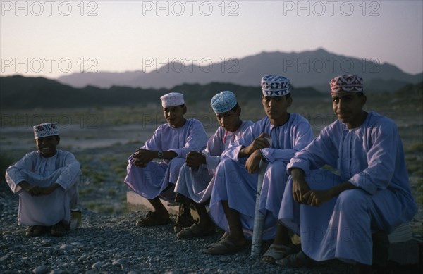OMAN, Children, "Schoolboys wearing traditional white dishdashas and brimless, embroidered circular hats sitting outside near Bahla"