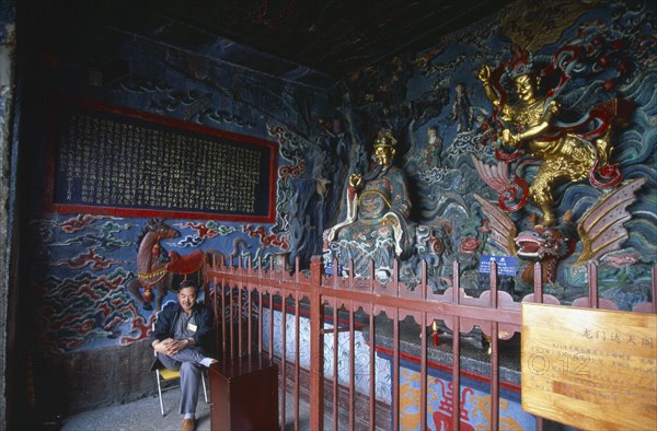 CHINA, Yunnan, Kunming , Interior view of the temple beside Dragon Gate in Western Hills with brightly painted walls and golden statuary and a man sat on a chair.