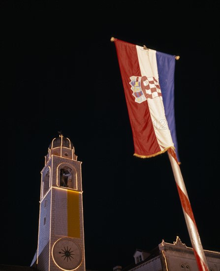 CROATIA, Dalmatia, Dubrovnik, View looking up at the Clocktower illuminated at night with Croatian flag in the foreground