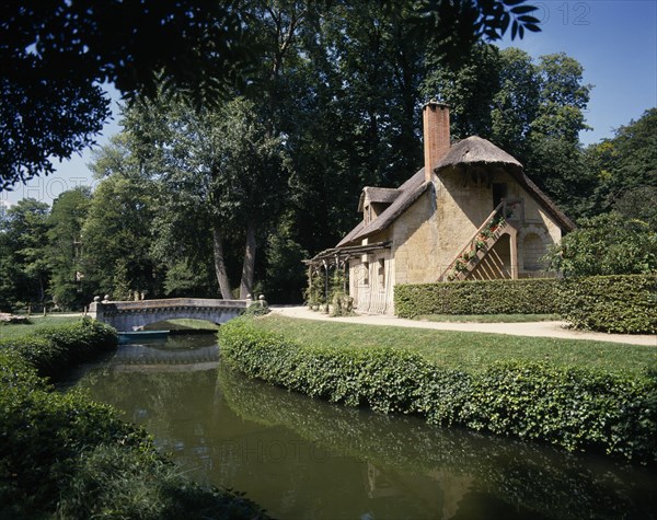 FRANCE, Ile de France, Paris, Versalilles Hameau de Trianon. View over river towards thatched cottage.