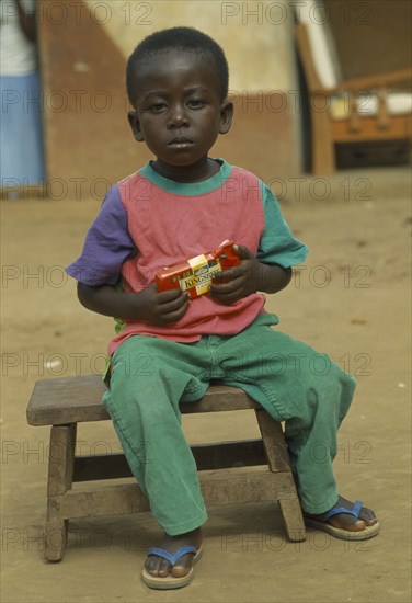 GHANA, Children, Portrait of young child sitting on wooden stool holding packet of chocolate.