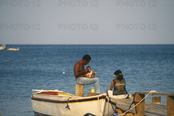 HONDURAS, Bay Islands, Roatan, Local couple with a baby sitting on wooden jetty at West End with small rowing boat tied to it.
