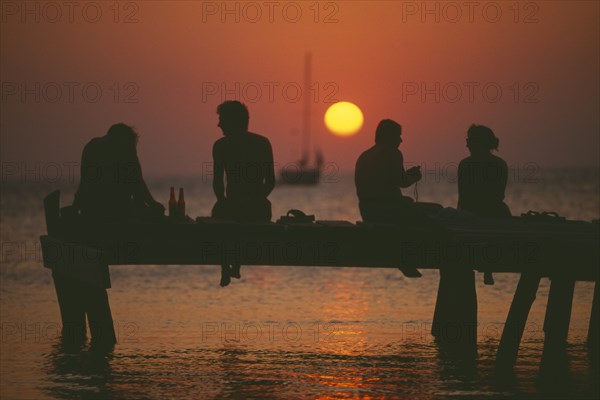 HONDURAS, Bay Islands, Roatan, Tourists sitting on jetty at West End silhouetted against warm orange sunset.