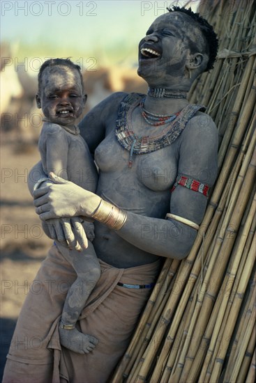 SUDAN, Mundari, "Agar Dinka woman decorated with dust, holding young son in her arms and laughing. Woman wearing marriage beads around her neck and white ivory bracelet on her arm."